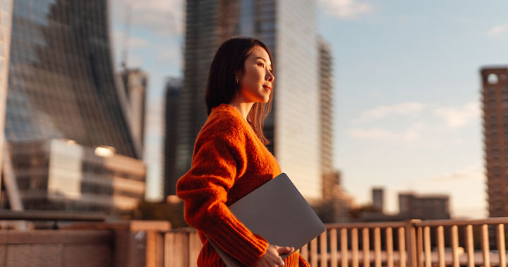 Thoughtful young Asian carrying laptop, looking at view in financial district at sunset. Concept of career achievement and investment planning.