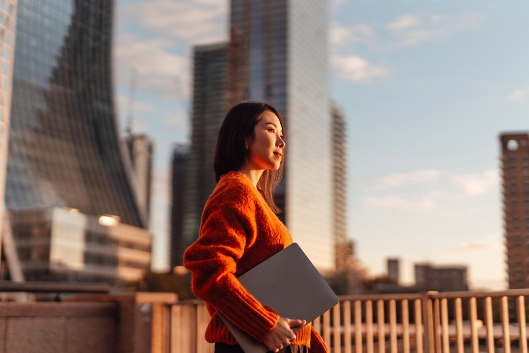 Thoughtful young Asian carrying laptop, looking at view in financial district at sunset. Concept of career achievement and investment planning.