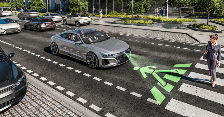 Image of a vehicle in a street scene in front of a pedestrian crossing, which a woman is currently crossing. The vehicle uses a video projector to project a pedestrian crossing icon directly onto the road in front of the vehicle.