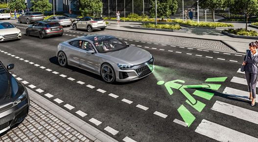 Image of a vehicle in a street scene in front of a pedestrian crossing, which a woman is currently crossing. The vehicle uses a video projector to project a pedestrian crossing icon directly onto the road in front of the vehicle.