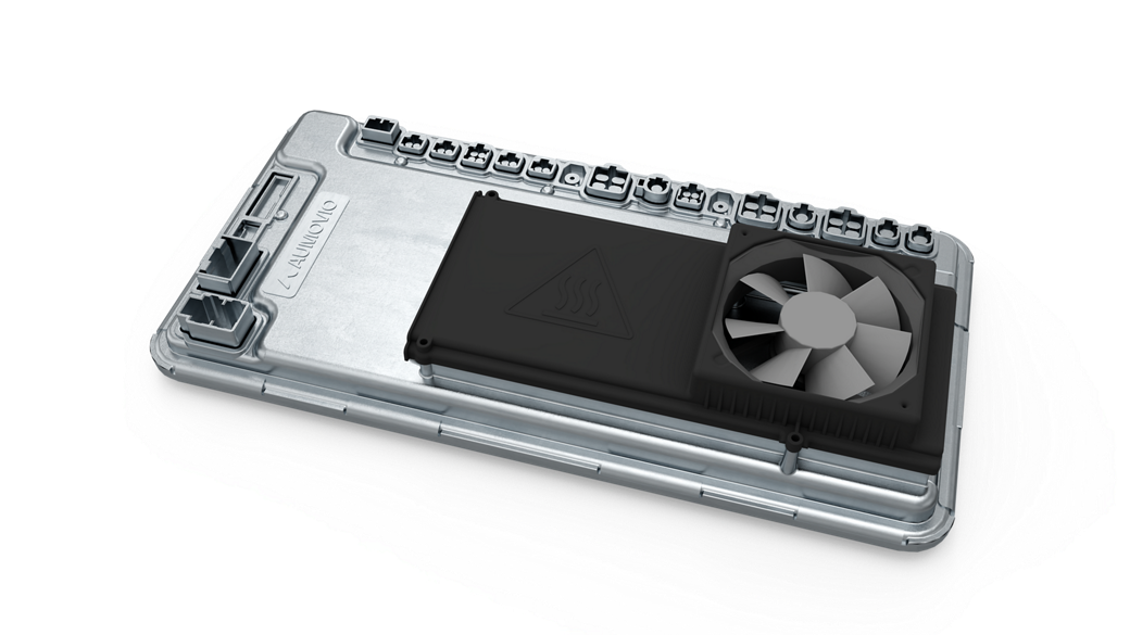 Close-up view of an automotive high-performance computer (HPC) unit featuring a metallic housing with multiple connector ports along the top edge. A black cooling module with a triangular heat warning symbol and an integrated fan is mounted on the surface, indicating active thermal management for high-performance processing in advanced vehicle systems.