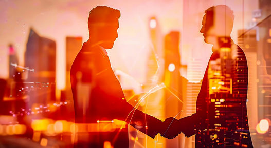 Two slightly transparent people shake hands in front of some illuminated skyscrapers in the city as the sun sets.