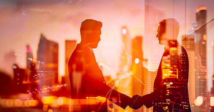 Two slightly transparent people shake hands in front of some illuminated skyscrapers in the city as the sun sets.