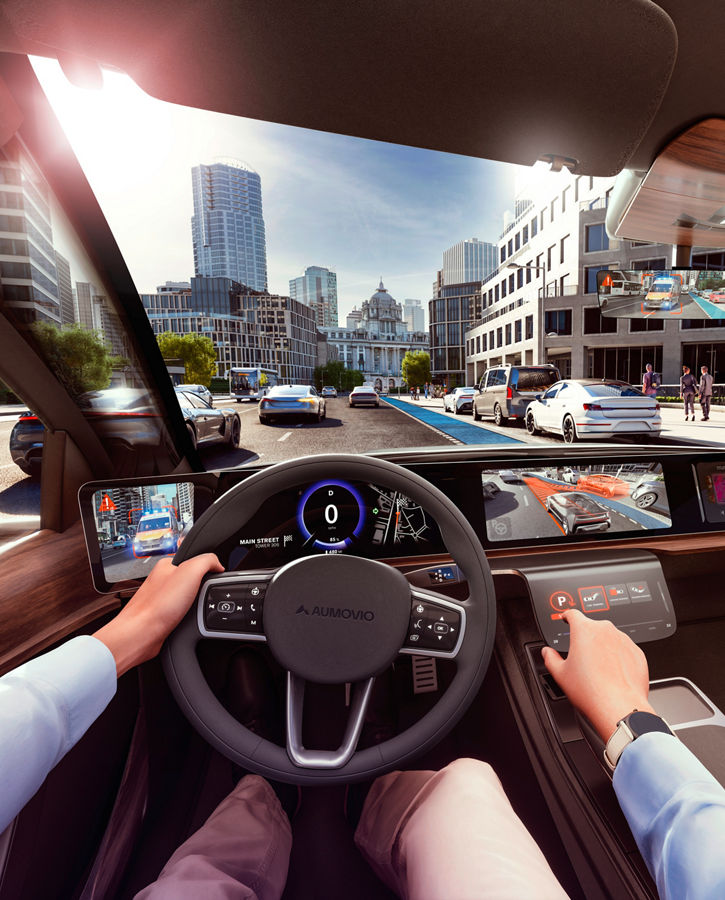 View through the windshield from inside the vehicle, from the driver's perspective over the dashboard to a traffic situation outside the vehicle. The external environment is displayed on several screens in the vehicle, including messages and warnings. AUMOVIO's state-of-the-art expertise in visualizing the vehicle and its surroundings is the result of many years of experience in the field of human vision.  Translated with DeepL.com (free version)