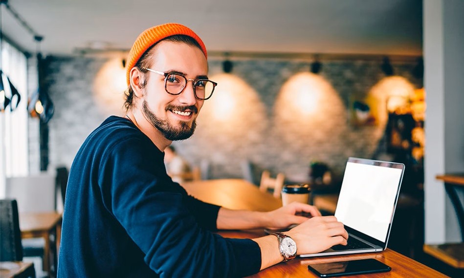 Eine Person sitzt an einem Holztisch in einem modernen Café und tippt auf einem Laptop, neben ihr liegen ein Smartphone und eine Kaffeetasse.
