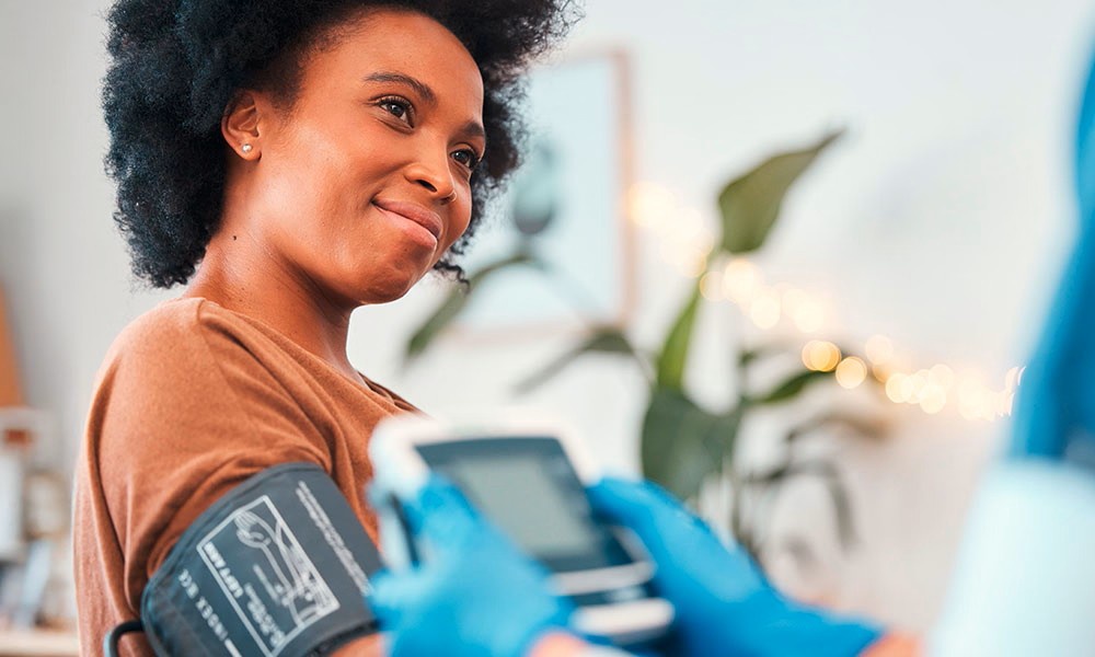 Medizinisches Fachpersonal mit blauen Handschuhen, das in einem gut beleuchteten Raum mit Pflanzen im Hintergrund ein digitales Blutdruckmessgerät bei einer Person anwendet.