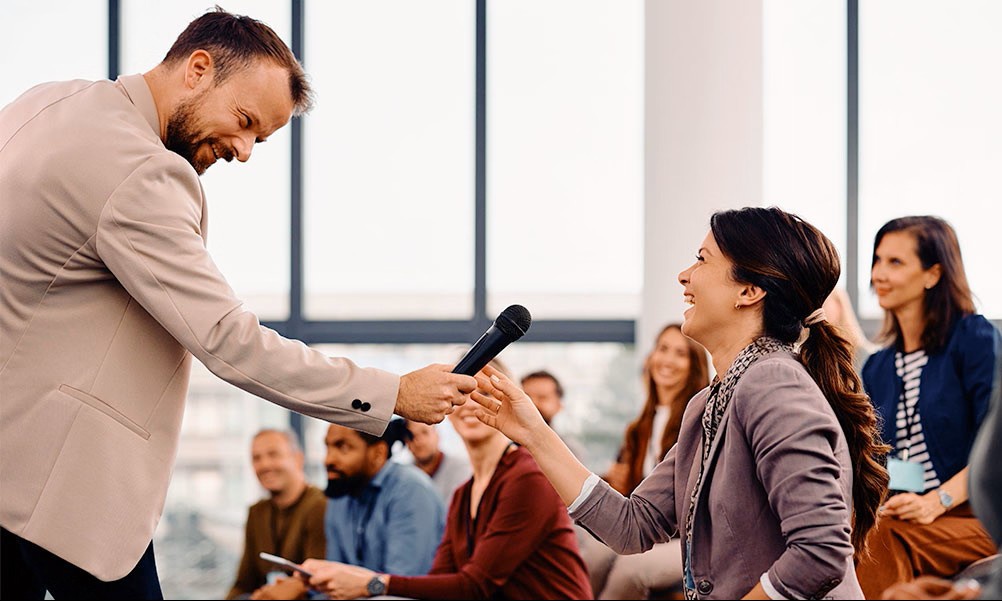 Person, die während einer interaktiven Sitzung in einem modernen Konferenzraum, in dem die Teilnehmer im Hintergrund sitzen, einem anderen Teilnehmer ein Mikrofon reicht.