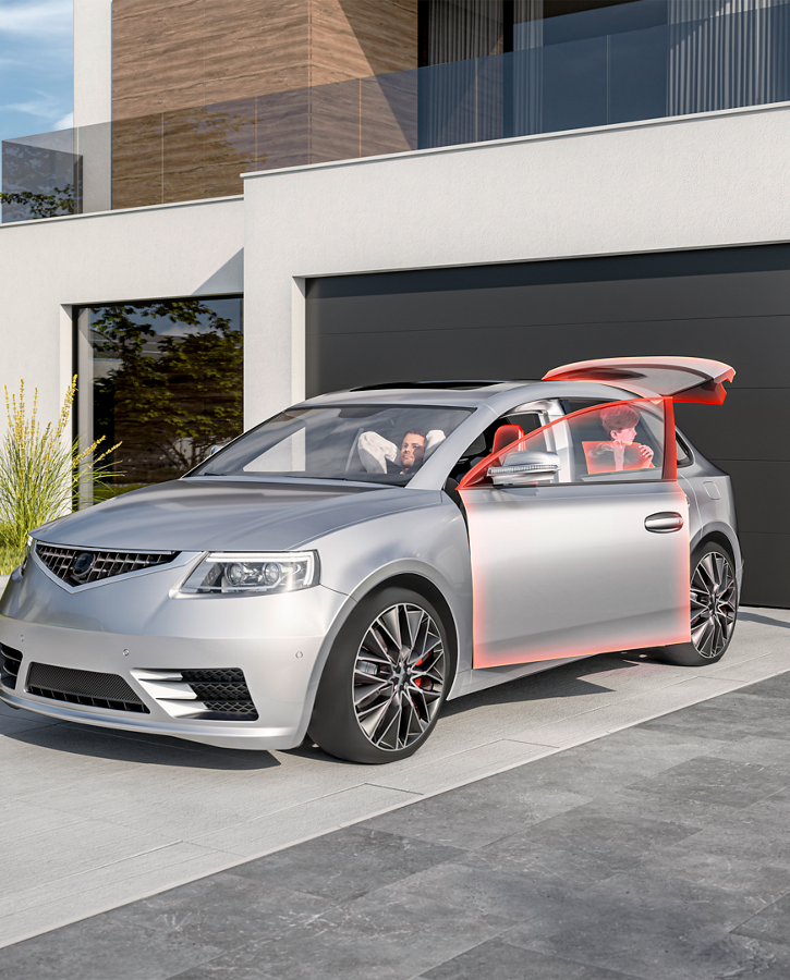 Modern silver sedan parked in front of a contemporary house with a large black garage door. The car’s front passenger door and rear hatch are open, highlighted in red, showcasing advanced body and actuator control features. A person stands nearby with a red suitcase, ready to load or unload luggage. The setting includes a clean gray driveway, green landscaped grass, and a stylish building with white walls, wooden accents, and large glass windows under bright daylight.