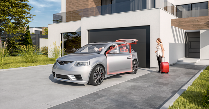 Modern silver sedan parked in front of a contemporary house with a large black garage door. The car’s front passenger door and rear hatch are open, highlighted in red, showcasing advanced body and actuator control features. A person stands nearby with a red suitcase, ready to load or unload luggage. The setting includes a clean gray driveway, green landscaped grass, and a stylish building with white walls, wooden accents, and large glass windows under bright daylight.