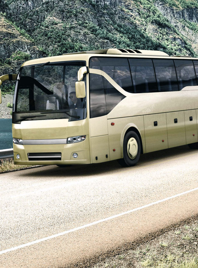 Modern beige coach bus driving on a scenic mountain road alongside a lake. The background features rocky hills and greenery, illustrating long-distance travel and commercial vehicle applications in a natural environment.