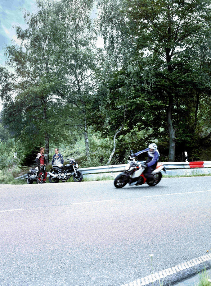 The picture shows a motorcyclist braking in a curve on a country road. In the background, two other motorcyclists and a quad bike can be seen. 