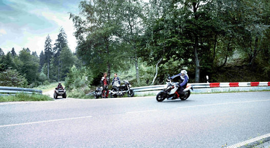 The picture shows a motorcyclist braking in a curve on a country road. In the background, two other motorcyclists and a quad bike can be seen. 