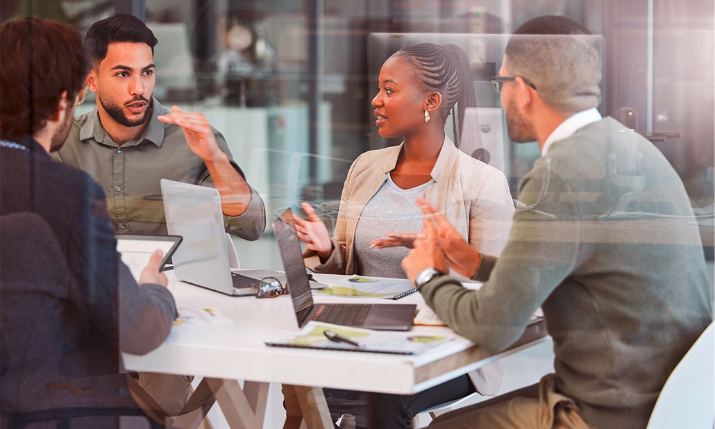 Vier Personen diskutieren an einem weißen Tisch in einem modernen Büro, auf dem Laptops und Dokumente verteilt sind.