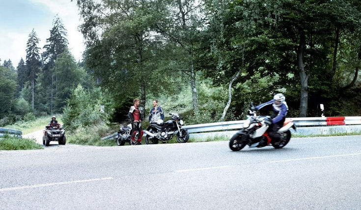 Motorcycles and an ATV parked on the side of a winding forest road with two riders standing next to their bikes. Another rider on a sport motorcycle is leaning into a curve, demonstrating dynamic cornering. The scene is surrounded by lush green trees and guardrails with red and white markings, emphasizing a scenic rural setting.