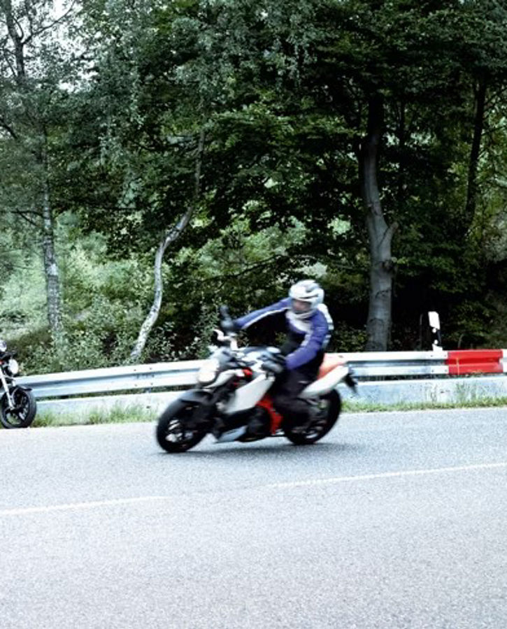 Motorcycles and an ATV parked on the side of a winding forest road with two riders standing next to their bikes. Another rider on a sport motorcycle is leaning into a curve, demonstrating dynamic cornering. The scene is surrounded by lush green trees and guardrails with red and white markings, emphasizing a scenic rural setting.