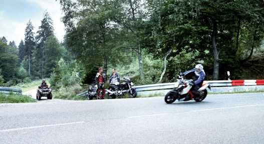 Motorcycles and an ATV parked on the side of a winding forest road with two riders standing next to their bikes. Another rider on a sport motorcycle is leaning into a curve, demonstrating dynamic cornering. The scene is surrounded by lush green trees and guardrails with red and white markings, emphasizing a scenic rural setting.