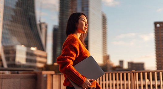 Thoughtful young Asian carrying laptop, looking at view in financial district at sunset. Concept of career achievement and investment planning.