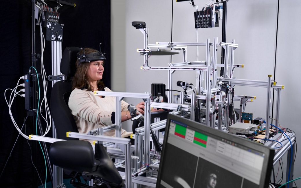 Laborumgebung mit einem Sitzgestell, auf dem eine Person in einem simulierten Fahrersitz sitzt, umgeben von einer Reihe von Sensoren und Metallrahmen. Ein Monitor im Vordergrund zeigt Echtzeit-Bilddaten an, die den Test- und Kalibrierungsprozess für fortschrittliche Sensorsysteme im Fahrzeuginnenraum veranschaulichen.