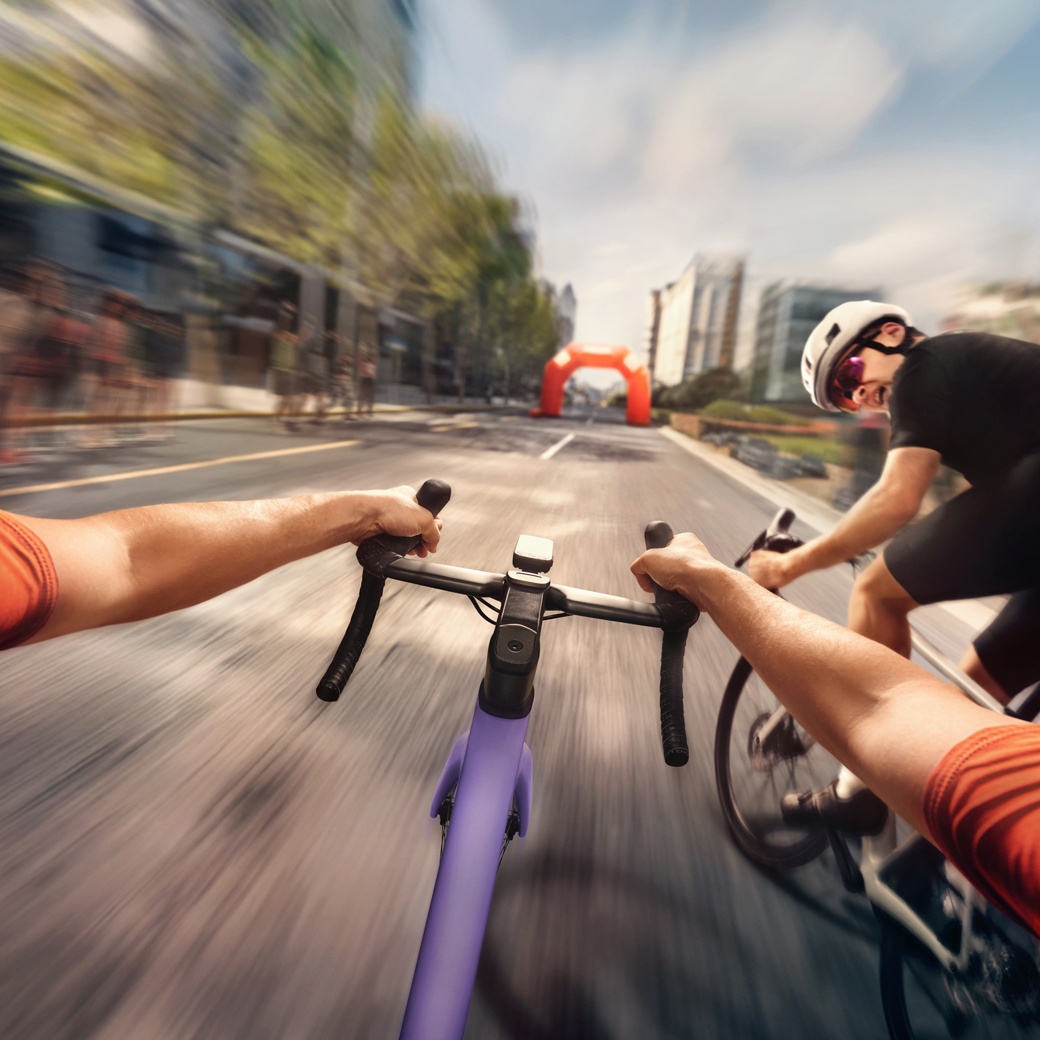   Dynamische Perspektive zweier Radfahrer, die auf einer Stadtstraße um die Wette fahren, wobei Bewegungsunschärfe die Geschwindigkeit betont und vor ihnen ein orangefarbener aufblasbarer Bogen zu sehen ist.