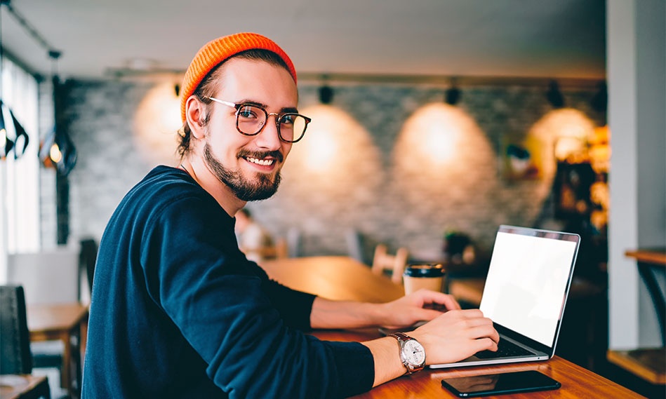 Eine Person sitzt an einem Holztisch in einem modernen Arbeitsbereich und tippt auf einem Laptop, neben ihr liegen ein Smartphone und eine Kaffeetasse.