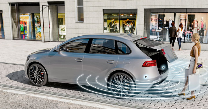 The image shows AUMOVIO's Kick Sensing software solution. A woman carrying bags in both hands opens the trunk by placing her foot under the rear of the vehicle.