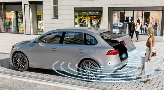 The image shows AUMOVIO's Kick Sensing software solution. A woman carrying bags in both hands opens the trunk by placing her foot under the rear of the vehicle.