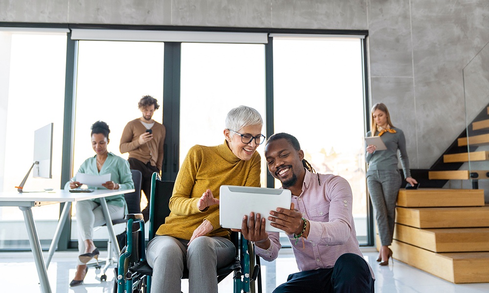 Two colleagues, one in a wheelchair, smile and look at a tablet together in a modern office. 