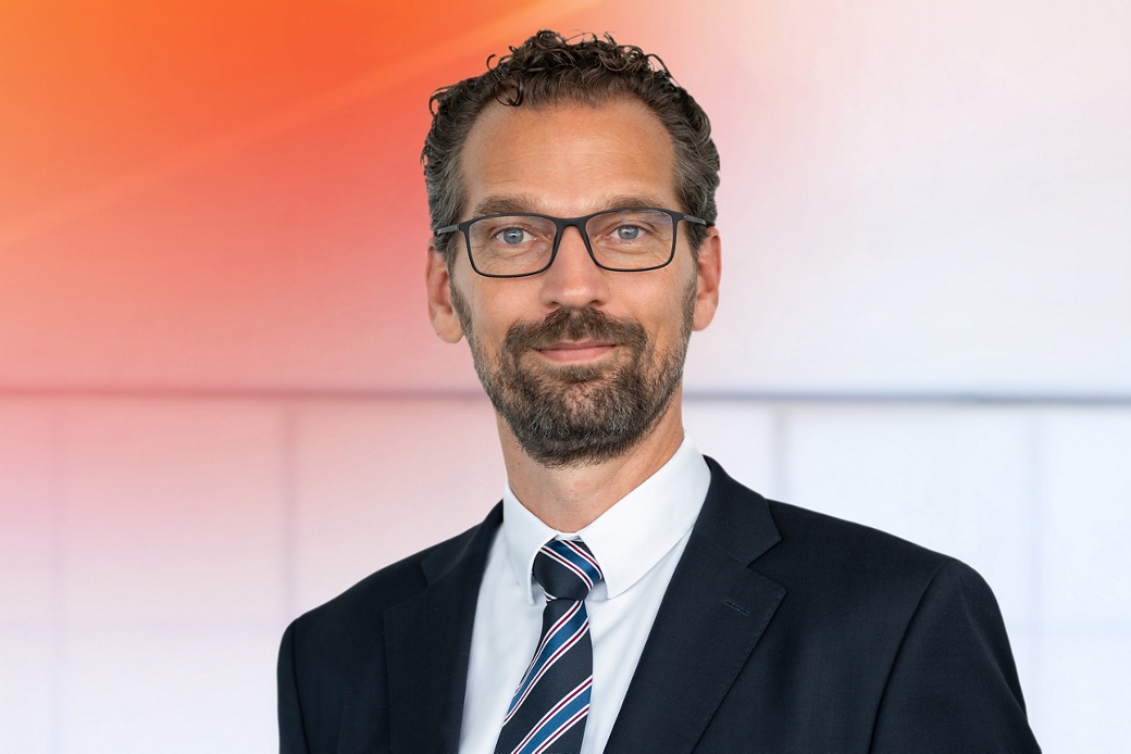 Dr. Boris Mergell in einem formellen dunklen Business-Anzug mit weißem Hemd und diagonaler gestreifter Krawatte in Blau-, Rot- und Weißtönen vor einem modernen Hintergrund mit Farbverlauf von Orange zu Weiß.