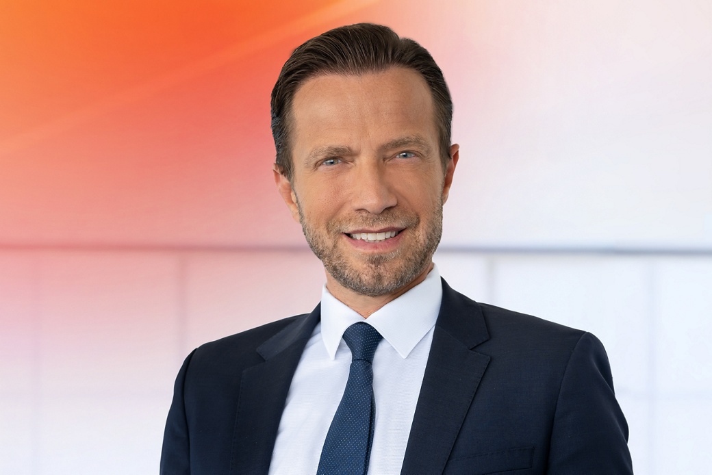 Ismail Dagli in einem formellen dunklen Business-Anzug mit weißem Hemd und einfarbiger dunkler Krawatte vor einem modernen Hintergrund mit Farbverlauf von Orange zu Weiß.