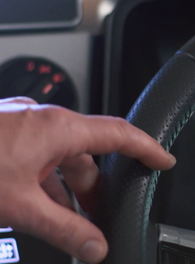 The image shows a truck or bus instrument cluster, with the driver's hand on the steering wheel visible in front of it. AUMOVIO driver's workstations for city buses or coaches offer a variety of options. With ergonomic designs and customizable features and various configurations to meet the needs of every driver.