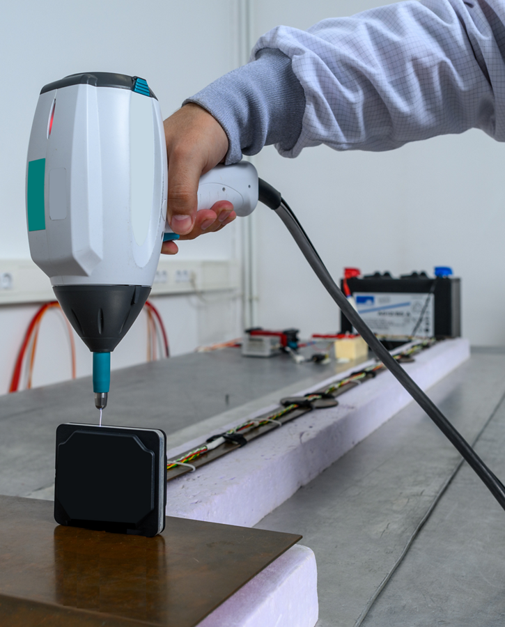 The picture shows a person using a testing device during electrical testing. AUMOVIO provides a globally aligned framework for validating the performance, reliability, and compliance of complex electronic systems across diverse industries.