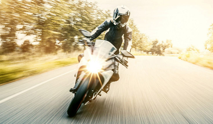 Motorcycle rider on a bright road with motion blur effect, wearing protective gear, with sunlight illuminating the scene, representing e-mobility for two-wheelers.