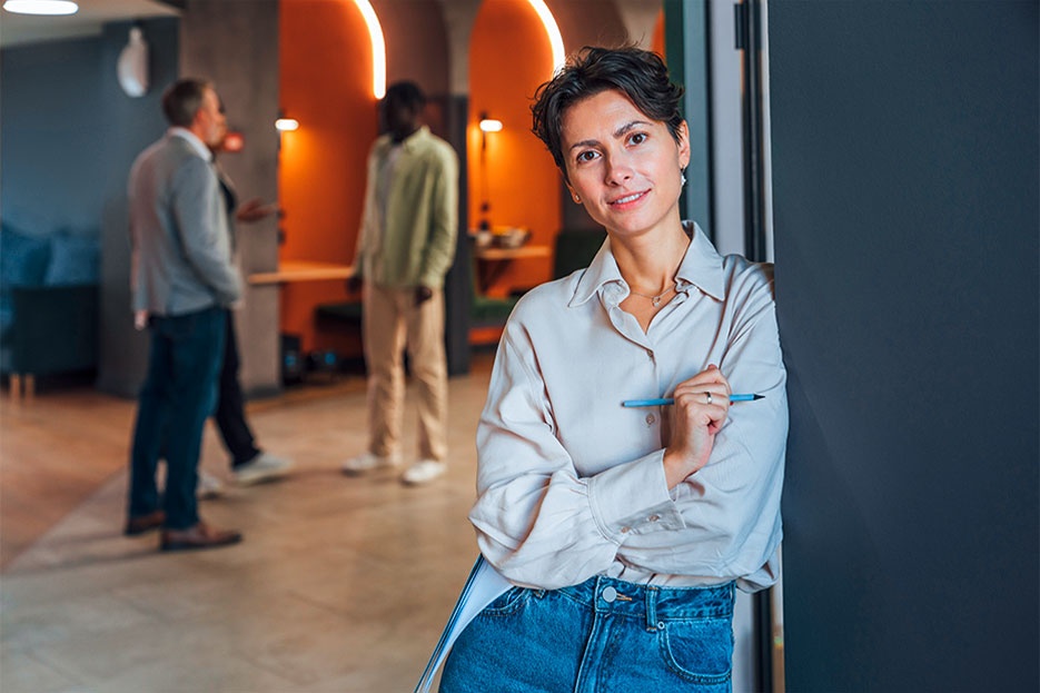 The equal opportunities image shows a woman in a break room or lounge area smiling at the viewer. Three other people, pupils or students, can be seen in the background. The image represents the idea that no matter what school qualifications you have, we at AUMOVIO have the right apprenticeship for you. Your personality, genuine interest, and motivation are our top priorities.