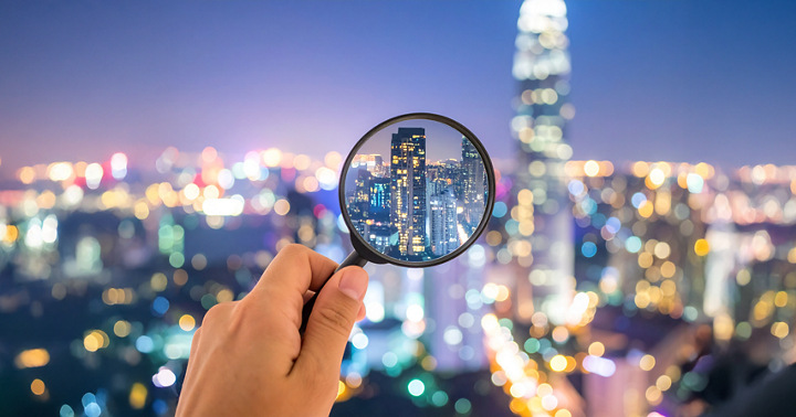 One hand holds a magnifying glass that enlarges a city with skyscrapers at night; in the background, many blurred lights of the city can be seen