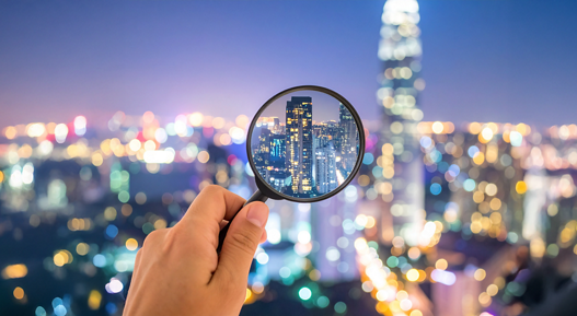 One hand holds a magnifying glass that enlarges a city with skyscrapers at night; in the background, many blurred lights of the city can be seen