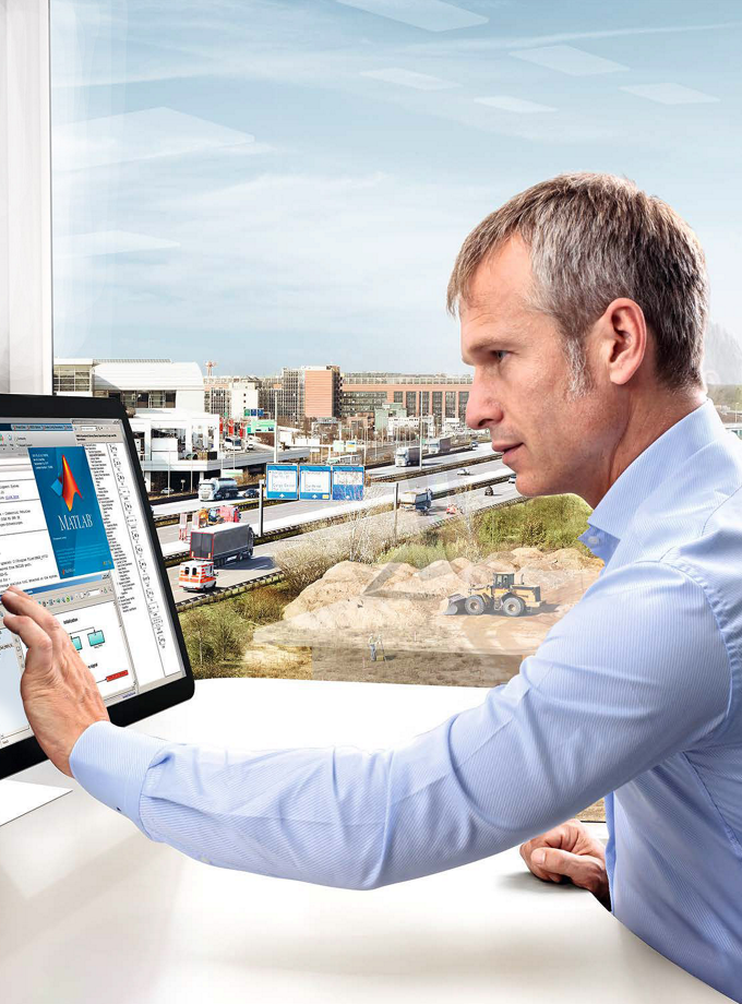 Desktop computer displaying automotive software interface for functional programming and configuration. The screen shows parameter settings and system diagrams, with a large window overlooking an industrial area and highway traffic in the background.