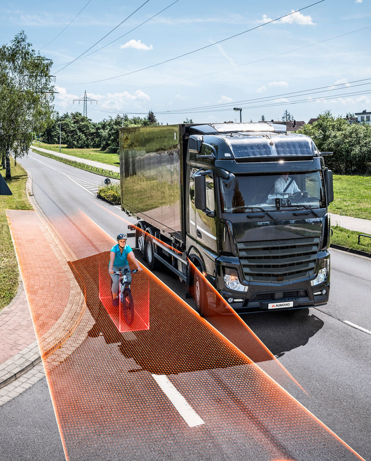 The image shows a cyclist riding right next to a truck on a bike path. The truck scans the area next to it—represented by orange lines and fields—to detect the cyclist and display them in the driver's rearview mirror.