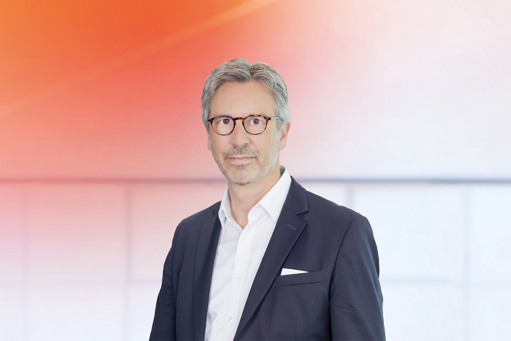 Ingo Holstein in einem formellen dunklen Business-Anzug mit weißem Hemd und Einstecktuch vor einem modernen Hintergrund mit Farbverlauf von Orange zu Weiß.