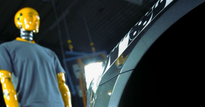 Image of a crash test dummy standing in front of a test vehicle in a proving ground facility