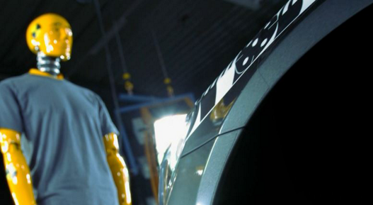 Image of a crash test dummy standing in front of a test vehicle in a proving ground facility