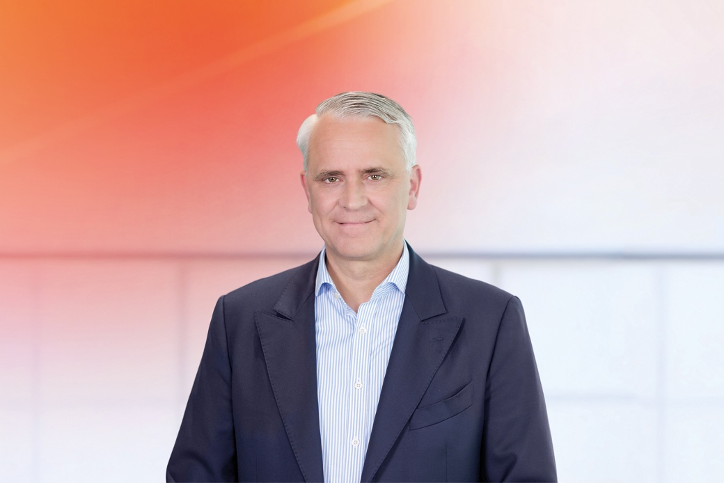 Philipp von Hirschheydt in einem formellen dunklen Business-Anzug mit hellblau gestreiftem Hemd vor einem modernen Hintergrund mit Farbverlauf von Orange zu Weiß.