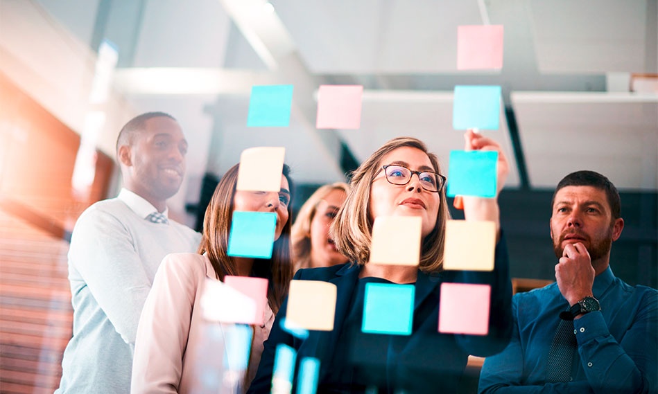 Eine Gruppe von Menschen, die in einer Büroumgebung zusammenarbeiten und hinter einer Glaswand stehen, die mit bunten Haftnotizen für die Planung und das Brainstorming bedeckt ist.