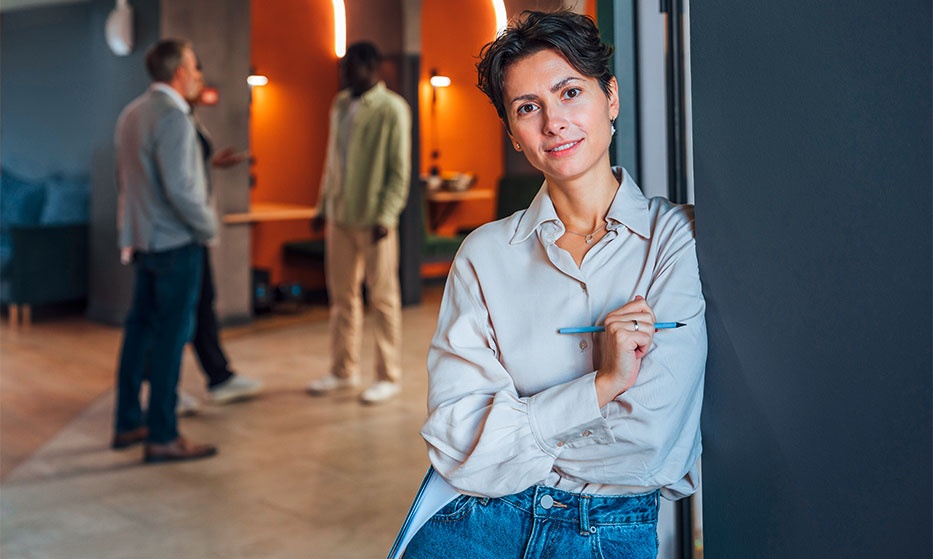 Eine Person lehnt lässig an einer Wand in einem modernen Büro, hält einen Stift in der Hand, während sich im Hintergrund zwei Personen unterhalten.
