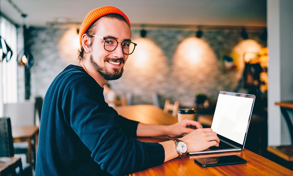 Eine Person sitzt an einem Holztisch in einem modernen Arbeitsbereich und tippt auf einem Laptop, neben ihr liegen ein Smartphone und eine Kaffeetasse.