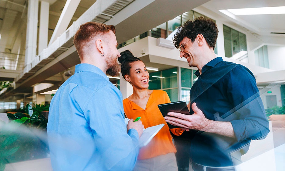 Drei Personen stehen zusammen in einer hellen, modernen Büroumgebung, halten ein Tablet und einen Notizblock in der Hand und unterhalten sich.