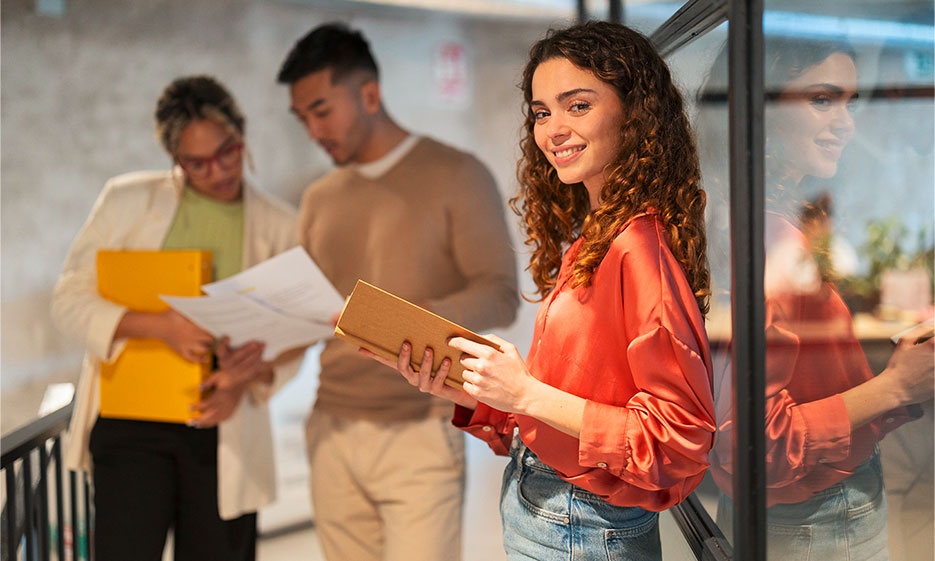 Eine Person steht mit einem Notizbuch in der Hand neben einer Glaswand in einem hellen Büroflur, im Hintergrund sind zwei Personen zu sehen, die Dokumente durchsehen.