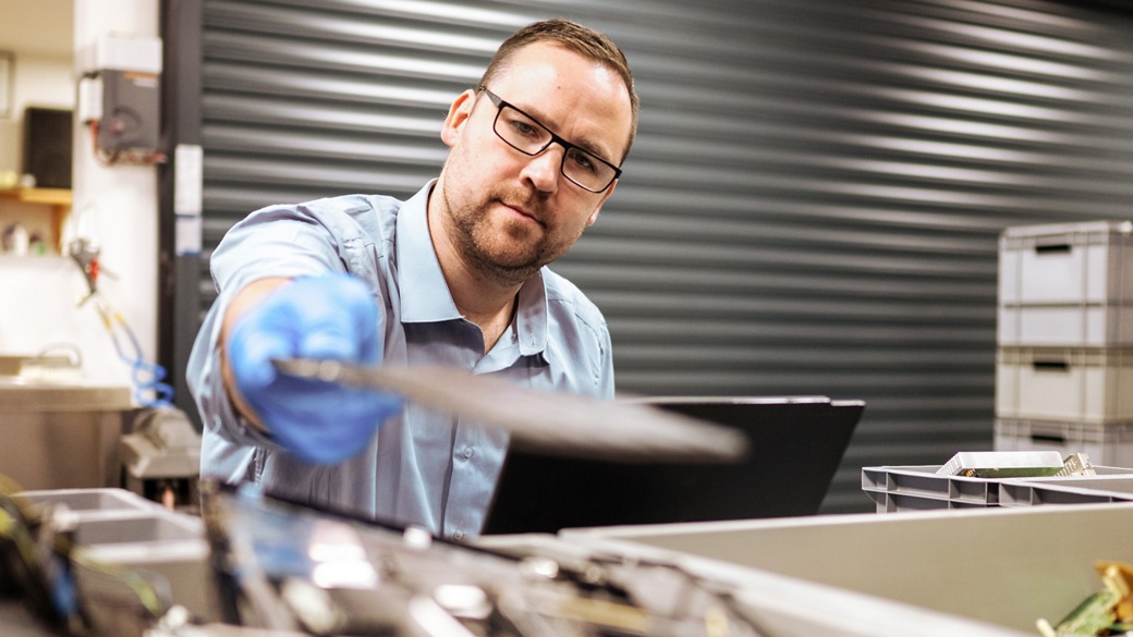 A man in a blue shirt and gloves is focused on his work in a workshop, carefully handling materials as part of a recycling or circular economy process. The setting appears to be a warehouse or industrial environment, emphasizing sustainability and resource management.