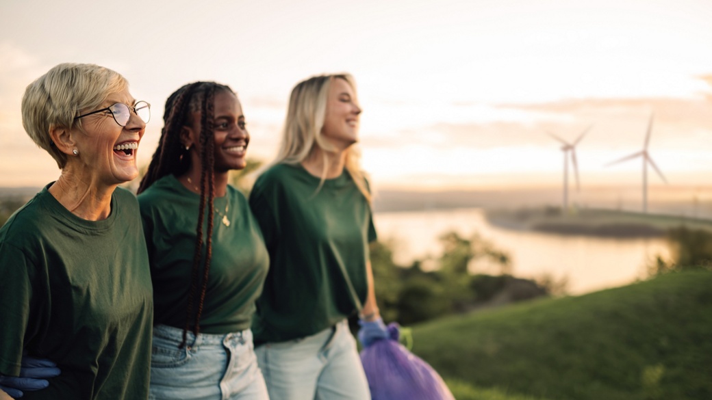 A diverse group of three people volunteering for environmental sustainability, smiling while working together outdoors at sunset. They are wearing green shirts and participating in a clean-up initiative, with wind turbines in the background.