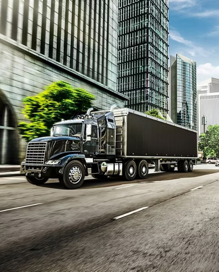 Large black heavy-duty truck driving on a multi-lane urban street surrounded by modern skyscrapers and glass buildings. The truck is hauling a long trailer, representing telematics and fleet management solutions for commercial vehicles. Bright daylight and city traffic create a professional logistics environment.