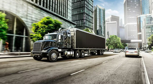Large black heavy-duty truck driving on a multi-lane urban street surrounded by modern skyscrapers and glass buildings. The truck is hauling a long trailer, representing telematics and fleet management solutions for commercial vehicles. Bright daylight and city traffic create a professional logistics environment.
